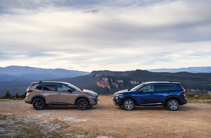 Zwei neue Nissan X‑Trail des Modelljahres 2026 stehen sich auf einer unbefestigten Straße gegenüber, im Hintergrund eine bergige Landschaft und bewölkter Himmel. Die Fahrzeuge präsentieren sich in kontrastierenden Farben: links der N-Trek in der Karosseriefarbe Baja Storm und rechts der Tekna in Universal Blue, beide mit markanten Felgendesigns und Dachreling, die den Outdoor-Charakter unterstreichen.