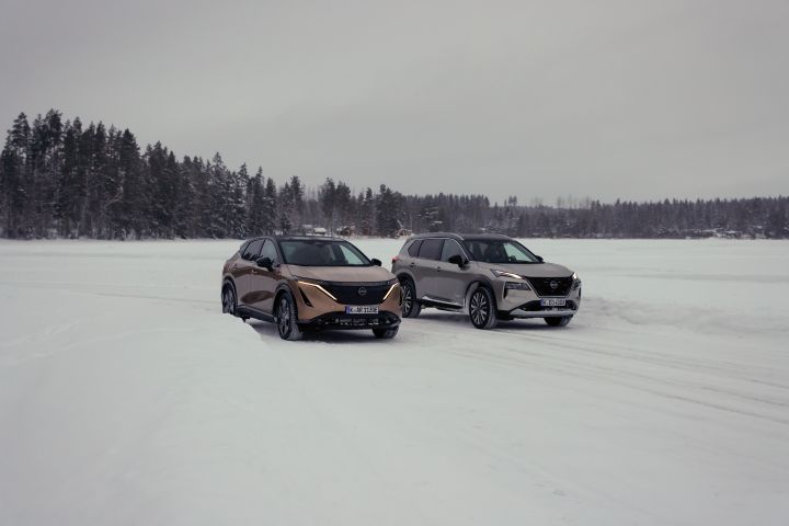 Elektroauto Nissan Ariya e‑4ORCE in Akatsuki Copper mit Dach und Außenspiegel in Pearl Black und silberner Nissan X‑Trail e‑Power e‑4ORCE fahren nebeneinander auf schneebedecktem Eissee mit verschneitem Wald im Hintergrund, 3/4 Front