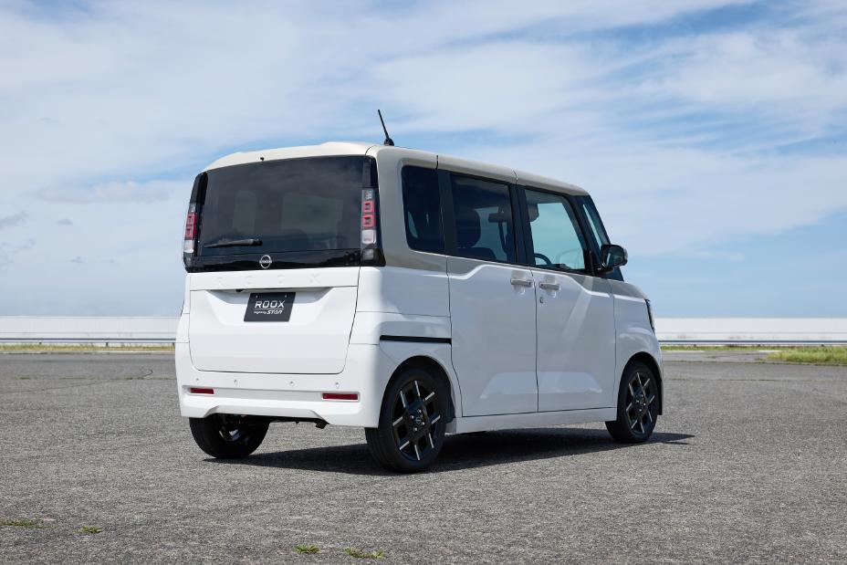 A white vehicle of the new Nissan Roox kei car is parked on a paved surface by the seaside, viewed from the rear. The background features the ocean, a visible horizon line, and a partly cloudy sky.