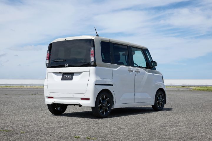 A white vehicle of the new Nissan Roox kei car is parked on a paved surface by the seaside, viewed from the rear. The background features the ocean, a visible horizon line, and a partly cloudy sky.