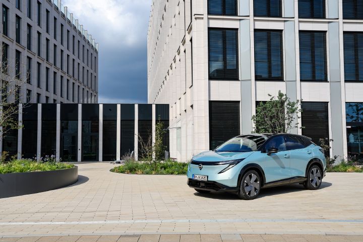 Auf einem hell gepflasterten Platz vor einem modernen Bürogebäude steht in der Sonne ein hellblauer Nissan Ariya, 3/4 Frontansicht