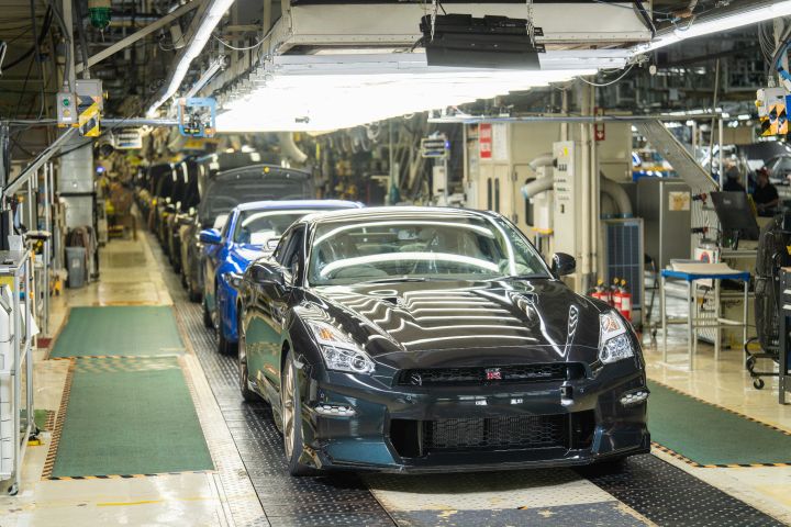 日産自動車 栃木工場の製造ラインにて、ミッドナイトパープルのR35 GT-Rが前景に写っており、後方には複数の車両が続いている。工場内は明るい照明に照らされ、周囲には自動車製造に使用される各種機械設備が配置されている。近代的な自動車生産工程の様子。