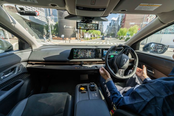 Ein Bild aus dem Innenraum eines Nissan Ariya, das dessen modernes und luxuriöses Interieur zeigt. Eine Person sitzt auf dem Fahrersitz des Rechtslenkers, ohne das Lenkrad festzuhalten. Im Hintergrund ist bei sonnigem Wetter eine städtische Straßenlandschaft mit Straßen und Gebäuden zu sehen.