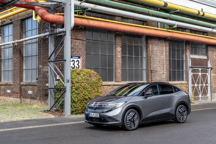 Vor der Wand eines Industriegebäudes steht ein grauer Nissan Leaf, 3/4 Frontaufnahme