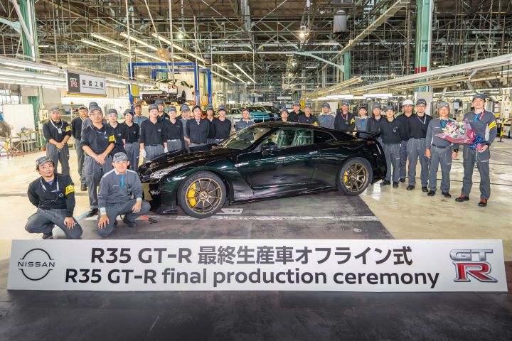 日産自動車 栃木工場の製造施設内にて、ミッドナイトパープルのR35 GT-Rを囲む複数の人物が写っている。車両は中央に配置され、前方には「R35 GT‑R 最終生産車オフライン式」と書かれた横断幕が掲げられている。背景には工場の設備が見え、R35 GT-Rの最終生産を記念する式典の様子。
