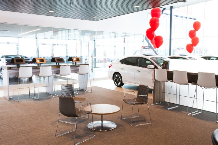 Salle d'exposition d'un concessionnaire automobile moderne avec une voiture blanche, des rangées de bureaux avec des ordinateurs et un coin salon avec des ballons rouges.