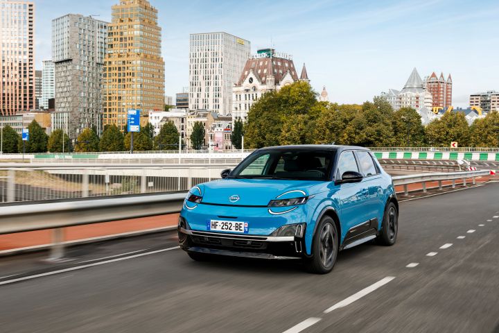Ein blauer Nissan Micra fährt im Sonnenschein über eine geschwungene Brücke, im Hintergrund sieht man verschiedene Gebäude aus dem Rotterdamer Stadtbild