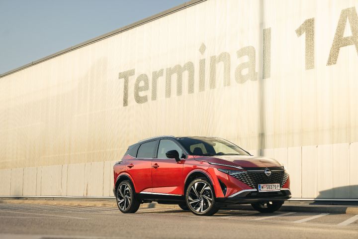 Blick von schräg vorne auf einen Nissan Qashqai in Fuji Sunset Red vor einem silbernen Gebäude mit der Aufschrift Terminal 1A