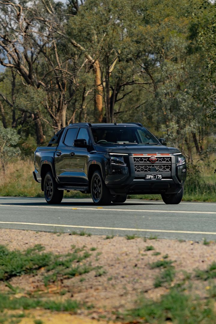 2026 Nissan Navara PRO-4X finished in Boulder Grey approaching on two‑lane bitumen with red grille accents, black wheels and side steps amid roadside gums.