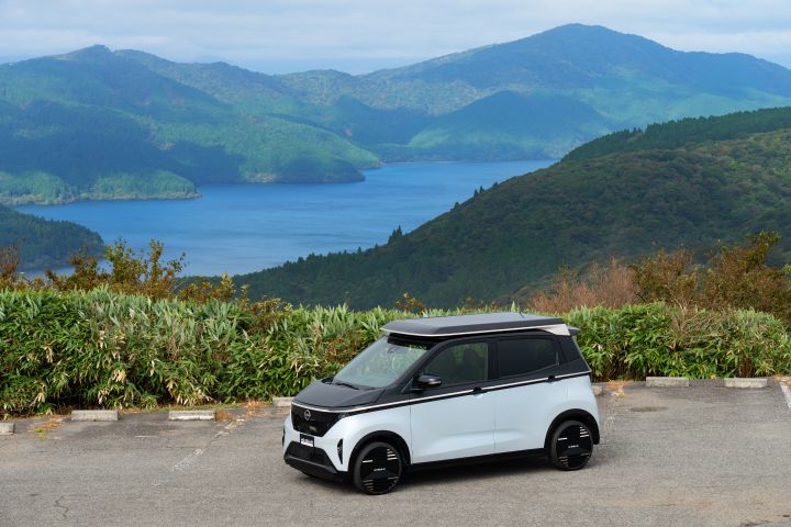 Against a backdrop of a lake and mountains, a light blue Nissan SAKURA is parked by the roadside, facing right. Green shrubs surround the area, and the sky is blue with scattered white clouds.