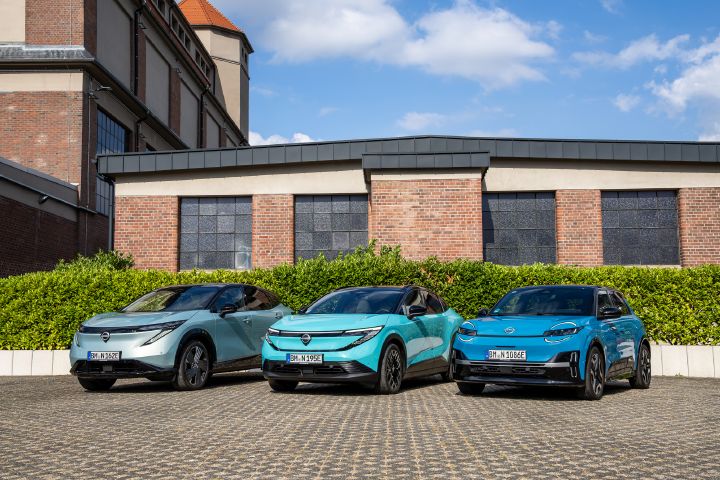 Vor einem Backsteingebäude und einer grünen Hecke stehen nebeneinander die drei Nissan EV-Modelle Nissan Ariya, Nissan Leaf und Nissan Micra, 3/4 Frontansicht aus leicht niedriger Position
