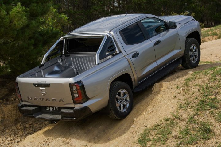 2026 Nissan Navara ST dual cab ute finished in Summit Silver shown from the rear-left driving along a dirt track with grassy embankments.