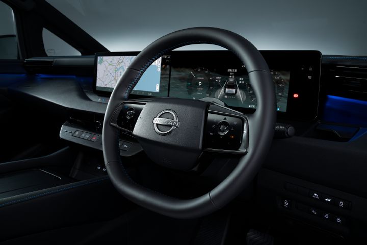 エルグランド AUTECHの運転席まわりを、運転席後方から前方に向かって撮影したインテリア画像。画面中央にはブラックのステアリングホイールが配置され、中央に日産エンブレムが見える。ステアリングの内側や縫い目には青色のステッチが施されており、AUTECHらしいスポーティかつ上質な印象を与えている。
ステアリングの奥には横長の大型ディスプレイが配置され、左側には地図を表示したナビゲーション画面、右側にはデジタルメーターが表示されている。メーター内には車両イラストや走行支援に関する表示、シフトポジション「P」などが確認できる。ダッシュボードはブラックを基調とし、ブルーのアンビエントライトが左右に伸び、高級感のある空間を演出している。
画面左側には助手席ドアとサイドウインドウの一部が見え、右側には各種スイッチ類が並ぶインストルメントパネルが配置されている。車内全体はブラックで統一され、青色の間接照明がアクセントとなっている。
