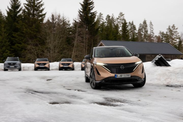Elektroauto Nissan Ariya e‑4ORCE in Akatsuki Copper mit Dach und Außenspiegel in Pearl Black fährt auf Schnee dynamisch um eine Kurve, im Hintergrund stehen weitere Nissan Autos