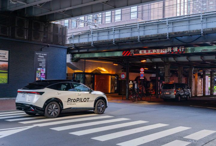 3/4 Heckansicht eines weißen Nissan Ariya, der auf einer Straße fährt, im Hintergrund ist eine Überführung mit Eisenbahnschienen oben und Geschäften unten zu sehen.