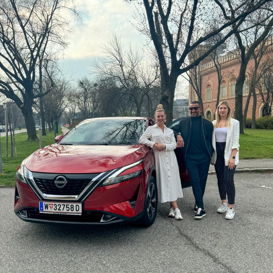 Fadi Merza steht lächelnd zwischen zwei ebenfalls lächelnden Frauen, alle drei stehen rechts neben einem roten Nissan Qashqai e‑POWER