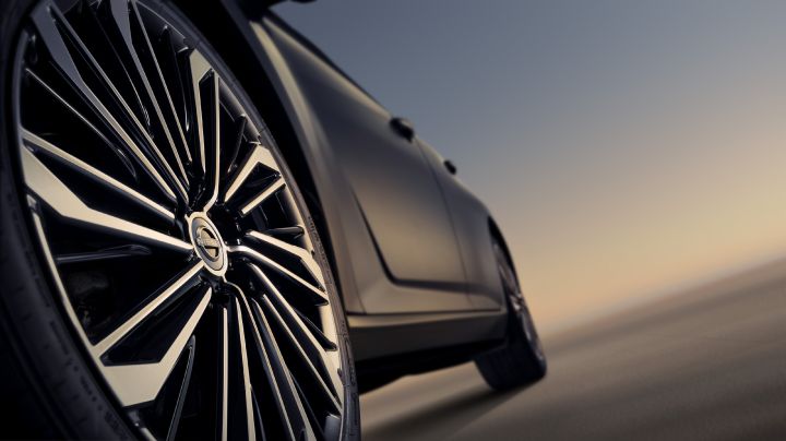 Angled close-up of a black Teana’s wheel, showcasing an intricate spoke design with a glossy finish. Part of the car body and a sunset background are visible.