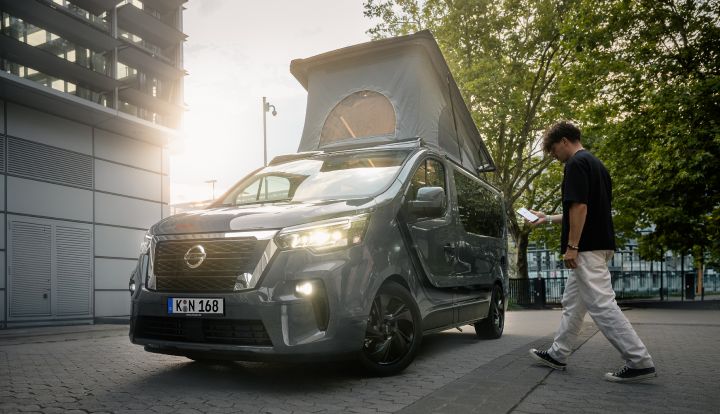 Auf einem Parkplatz steht ein grauer Nissan Primastar - Seaside by Dethleffs mit aufgestelltem Alkoven, ein junger Mann mit Smartphone in der Hand geht auf das Fahrzeug zu