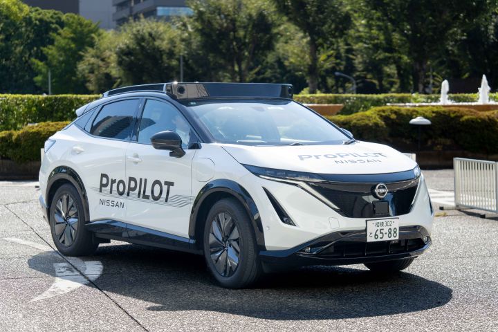 Ein weißer Nissan Ariya steht mit der Front nach links geparkt, an seiner Seite sind die Logos „ProPILOT” und „NISSAN” zu sehen. Das Fahrzeug steht auf einer asphaltierten Straße, im Hintergrund sind üppig grüne Bäume und Gebäude zu sehen.