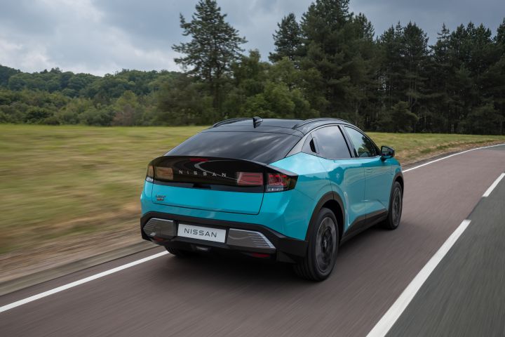 Photo dynamique 3/4 arrière de la nouvelle Nissan LEAF bleu turquoise traversant une route avec un espace vert et de grands arbres en arrière-plan