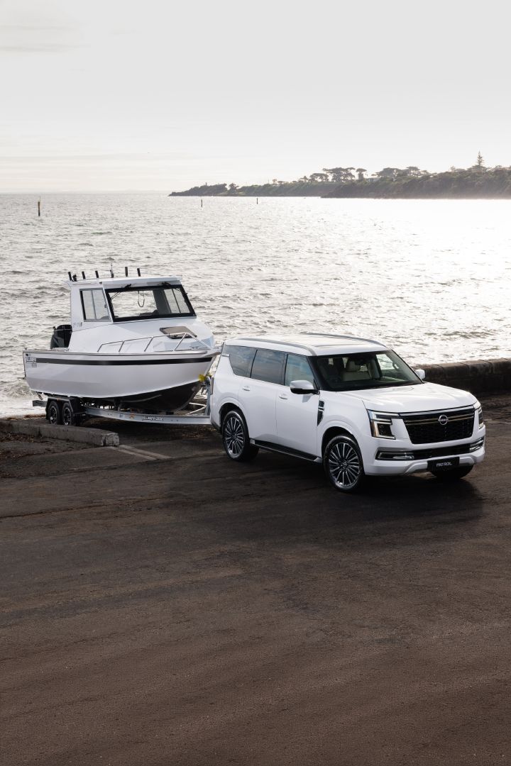 2026 Nissan Y63 Patrol finished in white, shown from an elevated front three-quarter exterior angle towing a large white boat on a trailer beside the ocean.