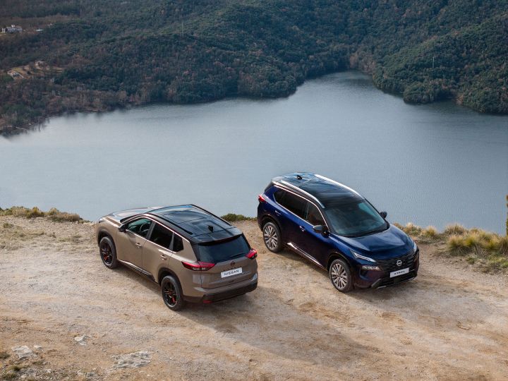 Zwei neue Nissan X‑Trail des Modelljahres 2026 stehen auf einer Felswand mit Blick auf ein großes Gewässer, das von bewaldeten Hügeln umgeben ist. Ein N-Trek SUV in der Karosseriefarbe Baja Storm mit schwarzen Akzenten und ein Tekna in der Karosseriefarbe Universal Blue sind so positioniert, dass ihr Außendesign und ihre Geländegängigkeit in einer natürlichen Umgebung zur Geltung kommen.