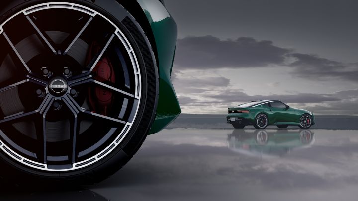 A close-up of the wheel of a metallic green Fairlady Z positioned on the left side of the image. The wheel is black with white accent lines and features a visible red brake caliper. In the distance, another green Fairlady Z appears small, angled toward the right-rear. The background is gray and misty, with a reflective surface and cloudy sky, creating a sleek and futuristic atmosphere.