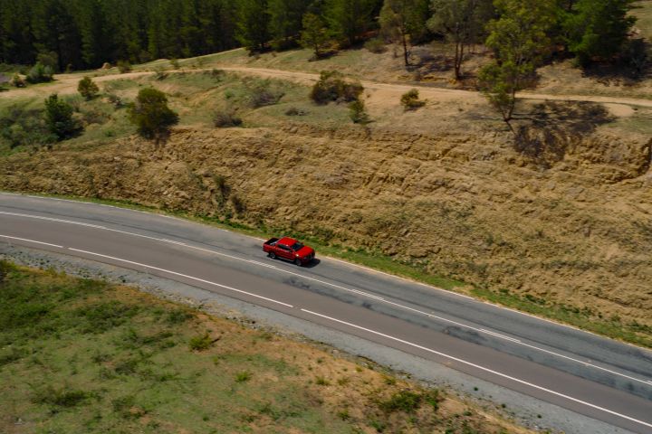 Overhead aerial shot of a 2026 Nissan Navara ST‑X dual cab painted in Outback Red, travelling on a sealed country road lined with brown and green grass.