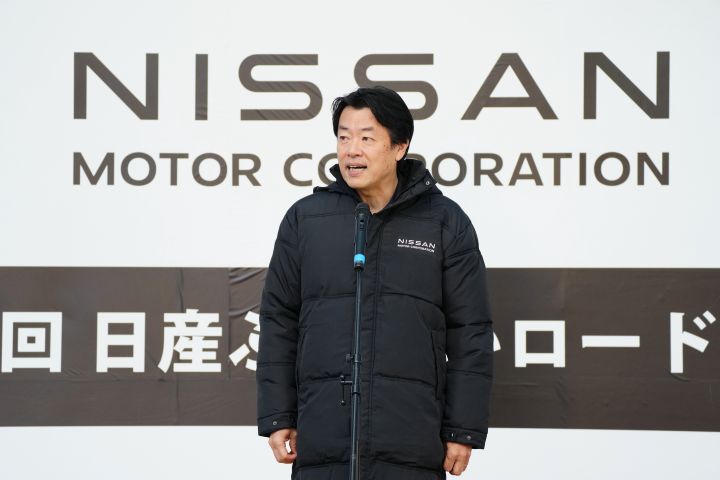 日産ロゴ入りの黒いコートを着た中畔EVPが舞台上でスピーチを行う様子