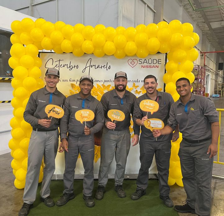 Quatro funcionários homens com uniforme da Nissan pousam para foto em frente a um painel do Setembro Amarelo com bolas amarelas no entorno.