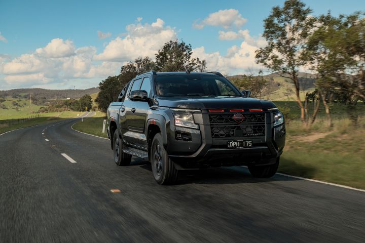 2026 Nissan Navara PRO-4X finished in Boulder Grey featured in a front-three-quarter cruising frame on a winding rural road with black side steps and distant rolling ranges.