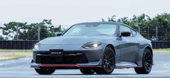 Gray Fairlady Z NISMO photographed from a front-right three-quarter angle on a rain-soaked race circuit, with the front of the vehicle facing toward the left side of the image. The car features a NISMO-specific front bumper, large grille, and red accent trim along the front splitter and side sills. Black alloy wheels and red brake calipers emphasize the performance-focused character of the model. Raindrops are visible across the bodywork and windows, while the wet track surface, tire barriers, safety fencing, and trees form the background. The image highlights the aggressive styling and dynamic presence of the Fairlady Z NISMO in rainy conditions.