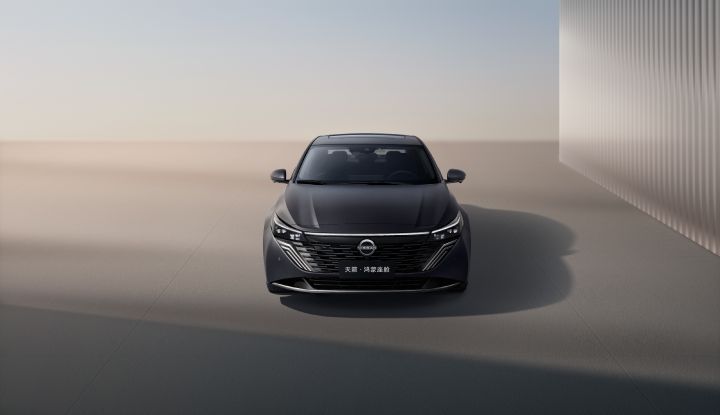 A black Teana parked in front of a building, facing left. The full side profile of the car is visible. The background shows a modern architectural design with light beige walls and vertical lines.