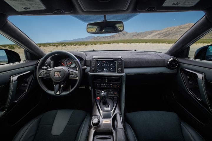 The steering wheel and dashboard inside the 2024 GT‑R T-spec Takumi, mountains are seen through the windshield .