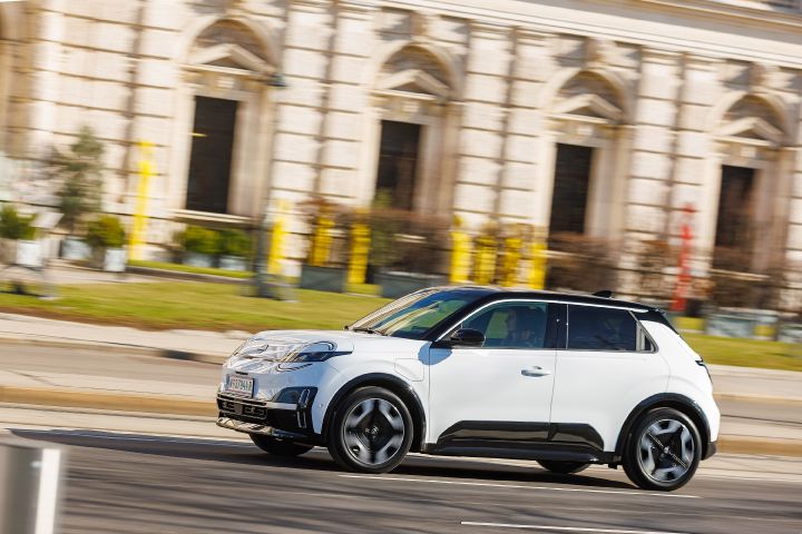7/8 Frontansicht eines weißen Nissan Micra mit schwarzem Kontrastdach, der zügig eine Straße entlang fährt mit gelben und roten Säulen vor Gebäuden im Hintergrund
