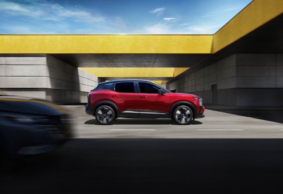 Side view of the Nissan KICKS in red, driving under the yellow bridge with car approaching from behind.