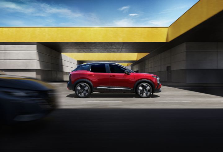 Side view of the Nissan KICKS in red, driving under the yellow bridge with car approaching from behind.