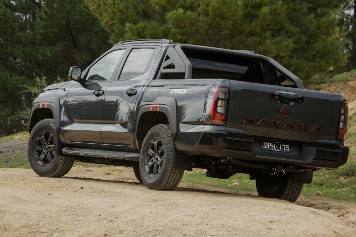 2026 Nissan Navara PRO-4X finished in Boulder Grey pictured rear three‑quarter on a sandy rise with NAVARA tailgate embossing, vertical tail‑lamps, towbar, sports bar and black wheels.