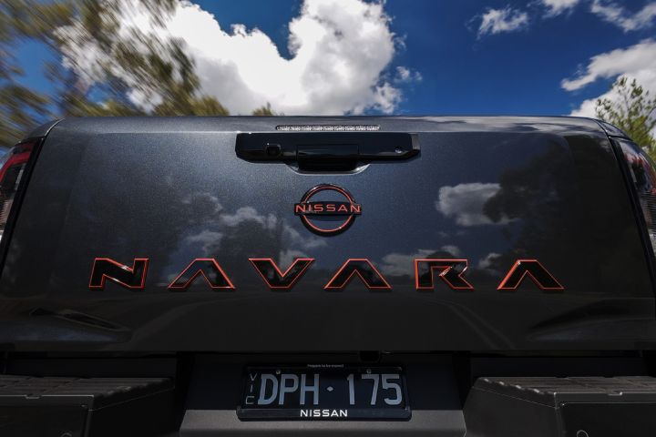 2026 Nissan Navara PRO-4X finished in Boulder Grey showing a tight tailgate crop with red-outlined NAVARA lettering, Nissan roundel, handle camera housing and cloud reflections.