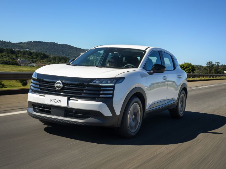 Nissan Kicks branco em movimento em uma rodovia, fotografado pela dianteira e lateral. Ao fundo, aparecem áreas verdes, colinas e céu azul.