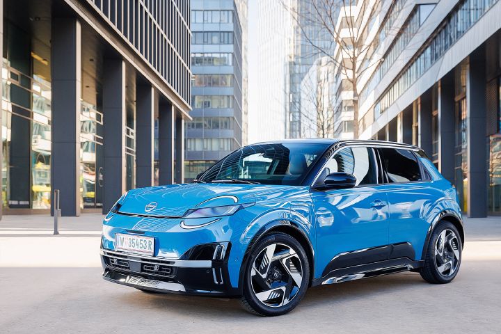 Blick von schräg vorne auf einen leuchtend blauen Nissan Micra mit schwarzem Kontrastdach, der zwischen mehreren Büro-Hochhäusern steht