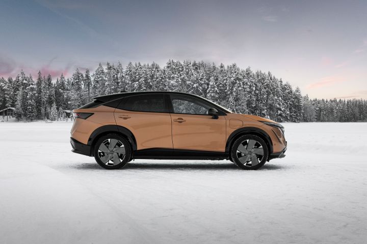 Elektroauto Nissan Ariya e‑4ORCE in Akatsuki Copper mit Dach und Außenspiegel in Pearl Black auf Eissee mit schneebedeckten Bäumen im Hintergrund, Seitenansicht