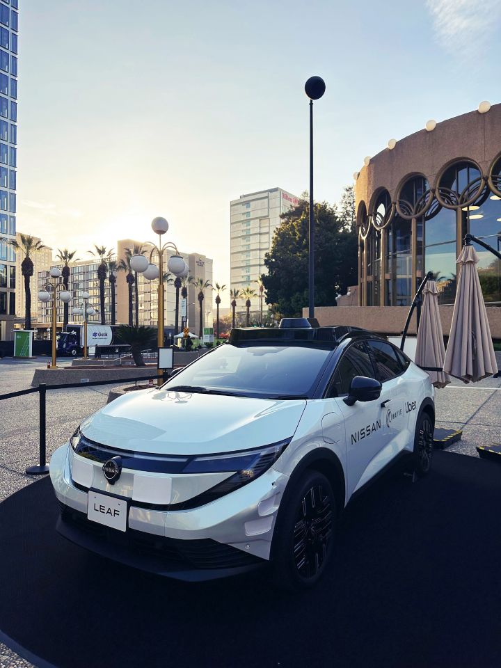A white robotaxi prototype based on the new Nissan LEAF is displayed outdoors in the early evening, shown from a front angled view. Tall buildings, a structure with round windows, and street trees appear in the background.