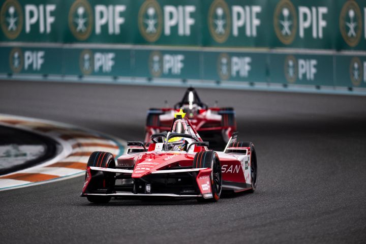 Oliver Rowland biegt mit seinem Nissan Formula E Rennwagen um eine Rechtskurve, direkt hinter ihm fährt ein zweites Formel E Auto, dahinter sind zwei grüne Werbebanden