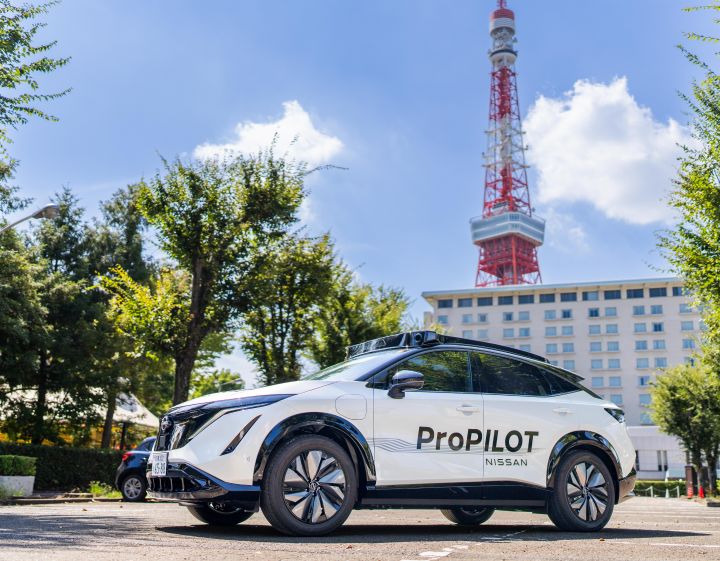 A white Nissan Ariya is parked facing left, with the "ProPILOT" logo visible on its side. In the background, a blue sky with white clouds stretches across, with the red and white Tokyo Tower standing prominently in the center. A large building is visible on the right, surrounded by lush green trees.