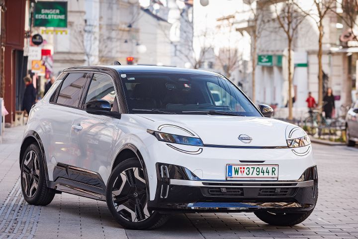 Auf einem gepflasterten Platz mit Geschäften im Hintergrund steht ein weißer Nissan Micra mit schwarzem Kontrastdach