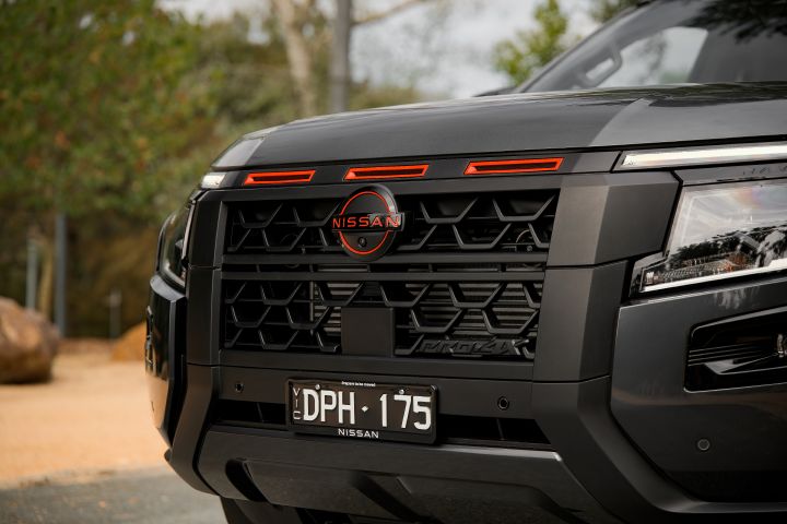 2026 Nissan Navara PRO-4X finished in Boulder Grey showing the front grille with red accents, Nissan badge, black mesh pattern, number plate and bumper.