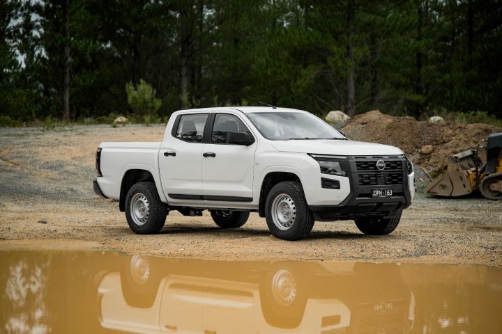 2026 Nissan Navara SL dual cab ute finished in Alpine White photographed from the front-left angle on a gravel worksite, with a muddy puddle reflection and forest backdrop
