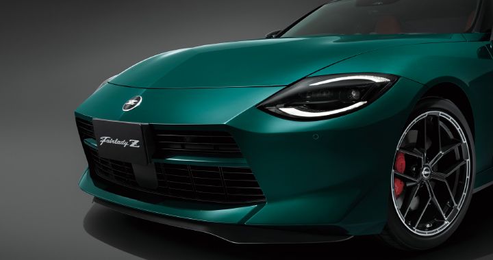 Close-up studio view of the front-right section of an Unryu Green Fairlady Z. The vehicle faces toward the left side of the image, highlighting the long hood, sharp LED headlamp, and large front grille. The front-right wheel features a black multi-spoke alloy design with a red brake caliper visible behind it, emphasizing the car’s sporty character. Set against a dark gray studio background, the rich Unryu Green paint finish and sculpted body surfaces are prominently showcased.