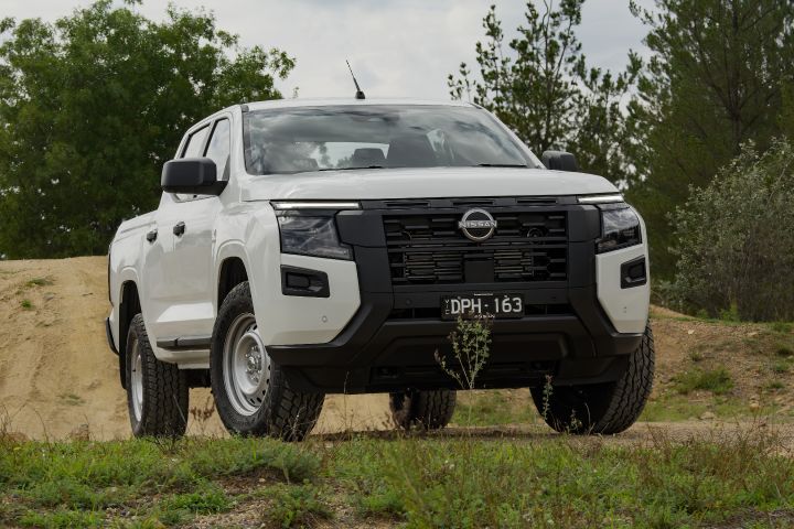 2026 Nissan Navara SL dual cab ute finished in Alpine White shown parked from a front three-quarter angle on grass and dirt near low scrub.